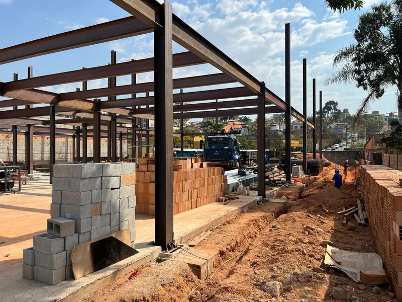 Pilares metálicos alinhados ao longo da obra, com valas de fundação e tijolos