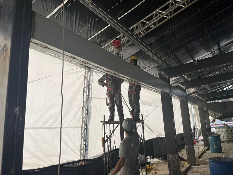 Montadores com cinto de segurança no andaime, junto às vigas escuras da estrutura