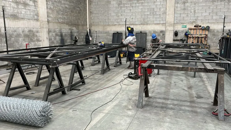 Soldadores montando os quadros tubulares dos portões sobre cavaletes, com o rolo de tela em primeiro plano